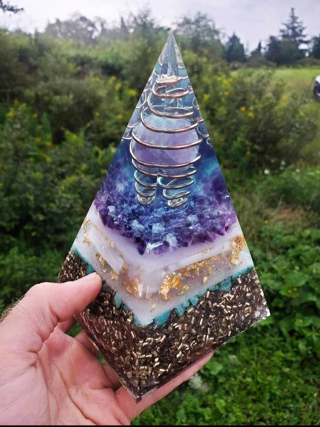 Nestled within the depths of this enchanting purple pyramid lies a harmonious blend of crystalline energies and sacred symbolism. Infused with the tranquil essence of amethyst, the purifying aura of selenite, and the soothing vibes of amazonite, it radiates serenity and balance. Brass and copper metals add their conducting touch, enhancing the energetic resonance within. Beneath its surface, the copper tensor ring and triskele symbolize the dance of cosmic forces, guiding the flow of energy with graceful precision. 